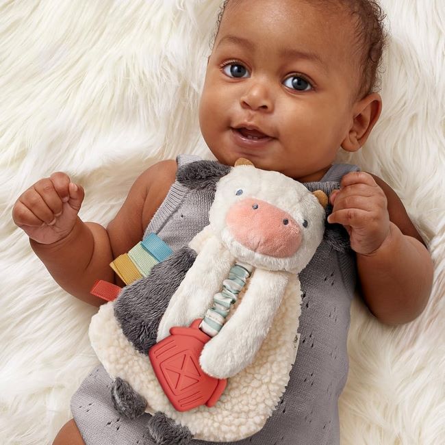 Baby cuddling Itzy Ritzy Itzy Lovey cow plush with teether, dangle arms, and crinkle textures.